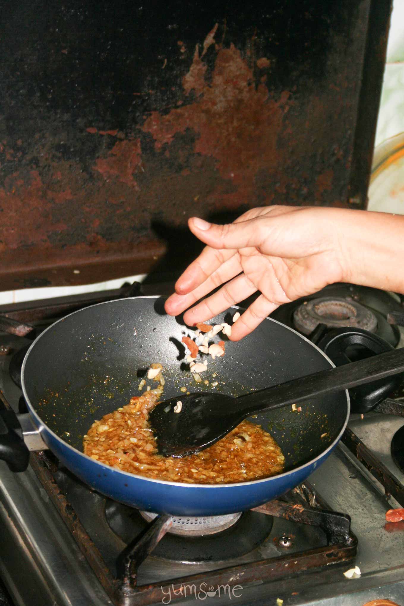 Usha's hand adding some ingredients to a kadai of spiced onions and garlic.