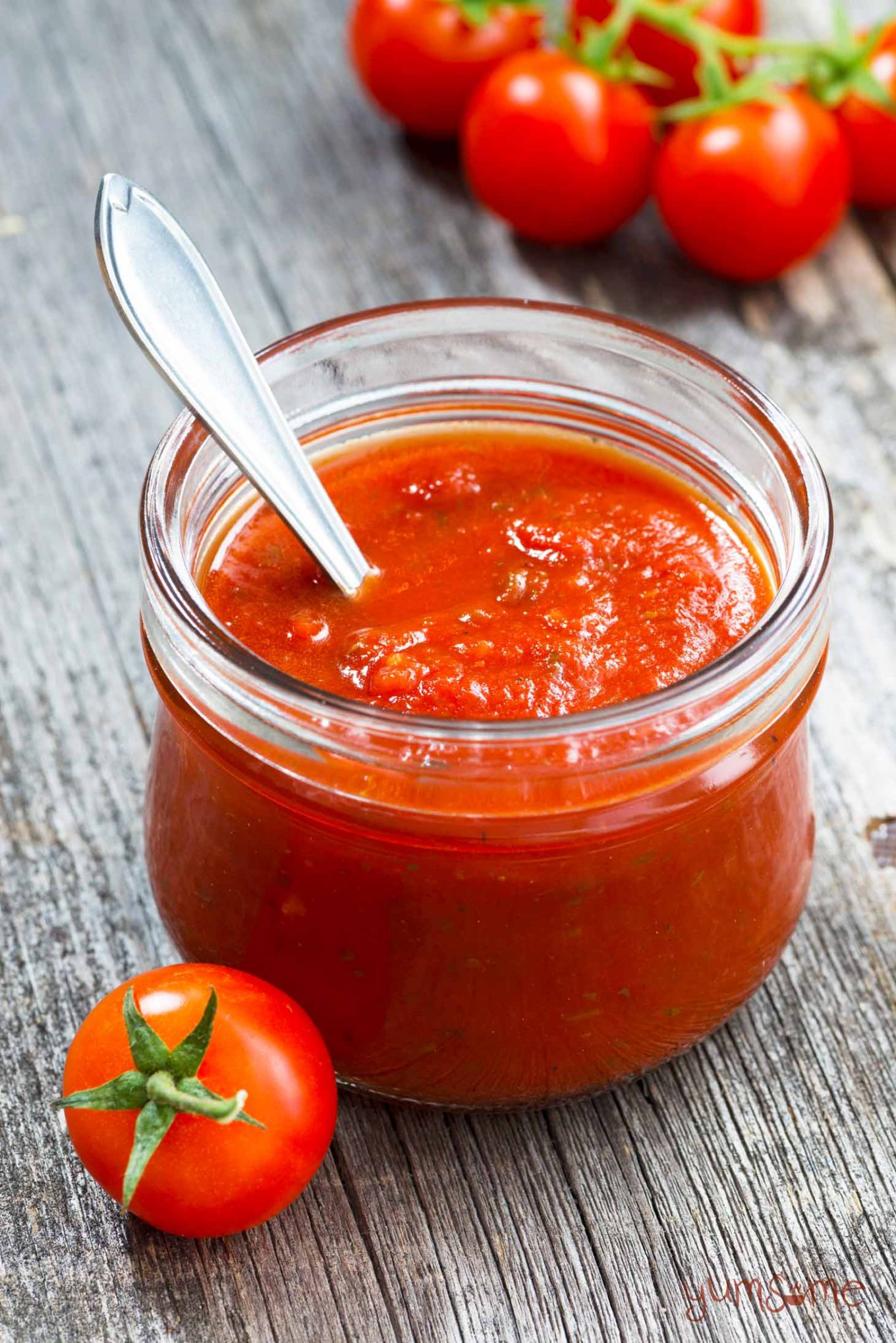 A spoon in a jar of classic Italian sauce, with some loose tomatoes scattered on a grey table.