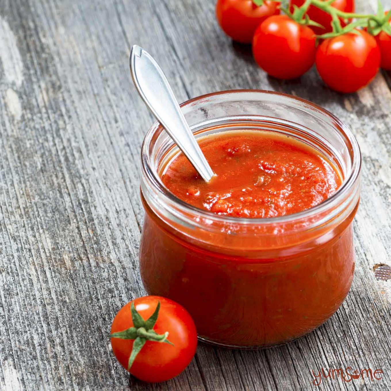 A small jar of classic Italian sauce with a spoon in it, on a grey table, with some loose tomatoes.