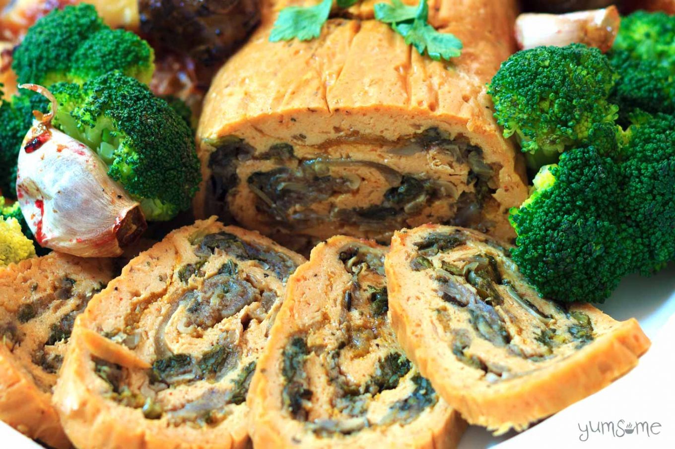 Closeup of a few slices of vegan stuffed seitan roast, with some steamed broccoli and roast garlic.