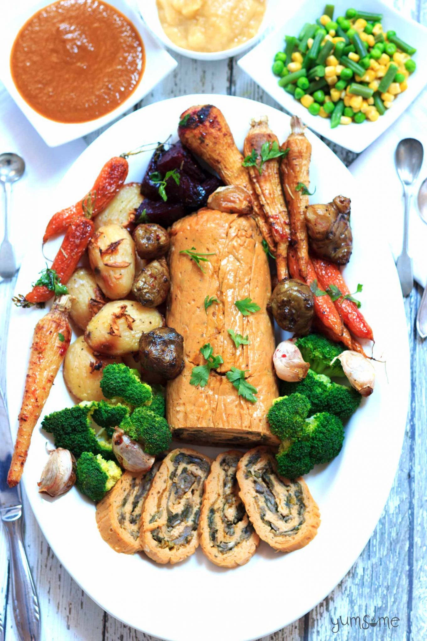 Overhead shot of a stuffed seitan roast on a platter, surrounded by roasted vegetables. Gravy, mashed potato, and steamed green beans are in white dishes in the background.