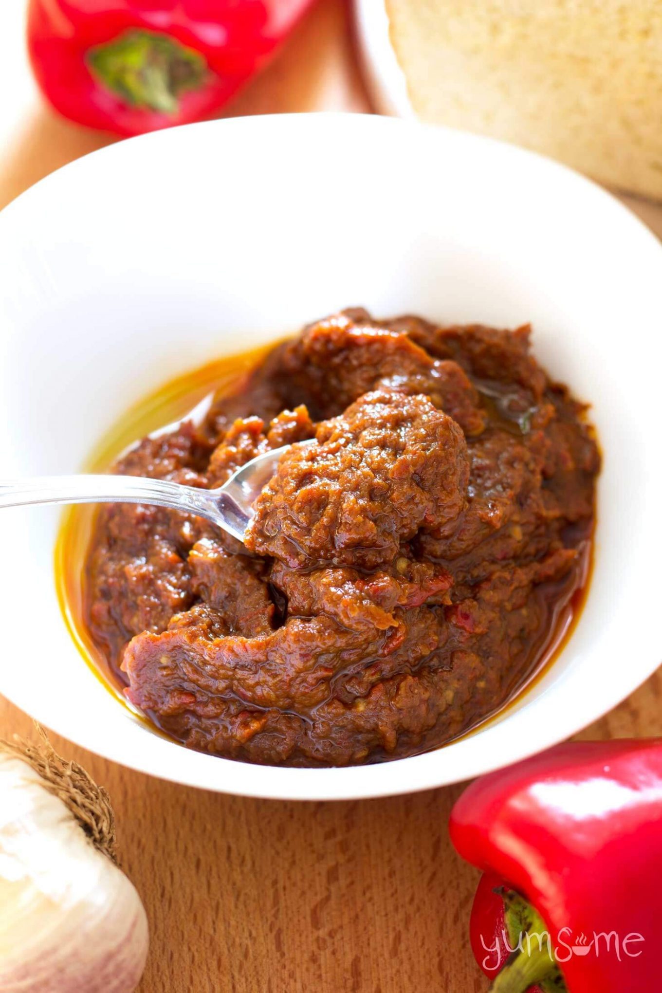 White bowl containing ajvar, on a wooden table, with two red peppers.