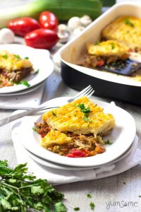 A white table set with several white dishes of vegan mushroom and buckwheat shepherds pie, with a large dish of shepherds pie in the background. There are also some fresh tomatoes, leeks, and parsley on the table.