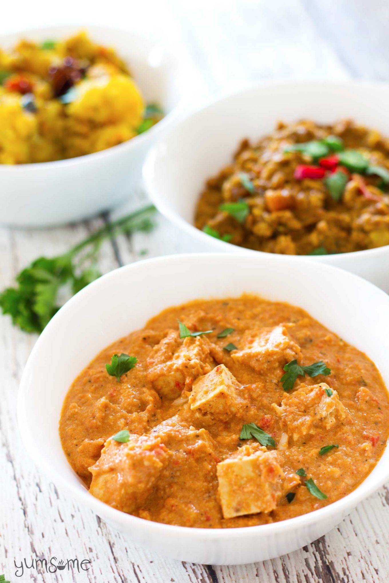 A bowl of vegan paneer butter masala on a white table, with bowls of vegan dal fry and aloo masala in the background.