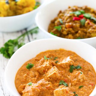 A bowl of vegan paneer butter masala on a white table, with bowls of vegan dal fry and aloo masala in the background.