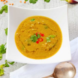 A white bowl containing vegan mulligatawny soup. A silver spoon is on a white napkin beside the bowl, and red chillies. lentils, and curry leaves are scattered in the background.