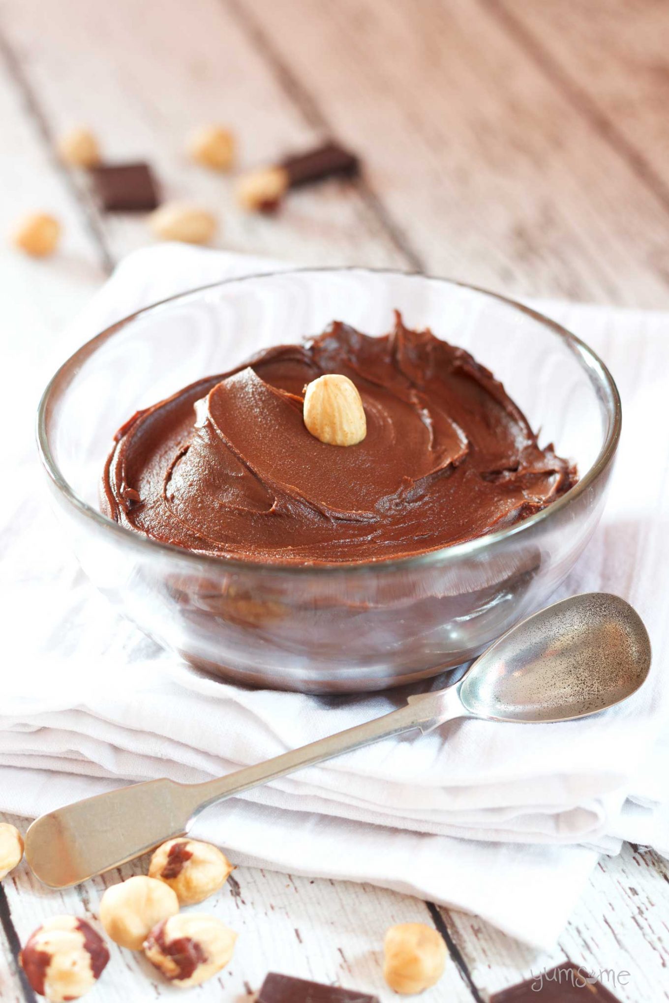 A bowl of vegan nutella with a hazelnut on top, on a white tea cloth with a silver spoon.