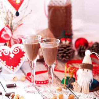 Two glasses and a bottle of vegan nutella cream liqueur on a white table, with hazelnuts and some pieces of chocolate in the foreground.