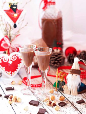 Two glasses and a bottle of vegan nutella cream liqueur on a white table, with hazelnuts and some pieces of chocolate in the foreground.