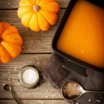 A bowl of pumpkin soup on a wooden table, and two pumpkins.