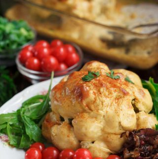 A whole cauliflower cheese on a whhite plate, surrounded by cherry tomatoes, green salad, and caramelised onions.