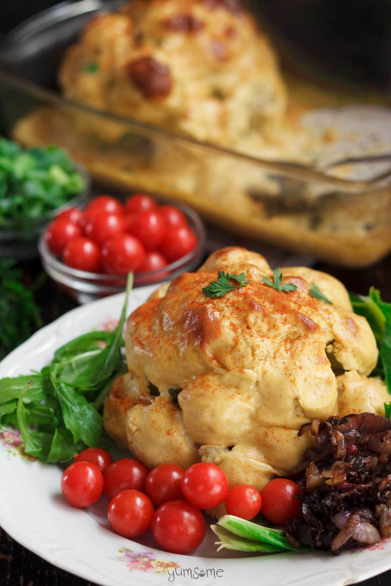 A whole cauliflower cheese on a whhite plate, surrounded by cherry tomatoes, green salad, and caramelised onions.