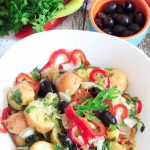 A white bowl of loaded potato salad, surrounded by herbs and vegetables on a white table.