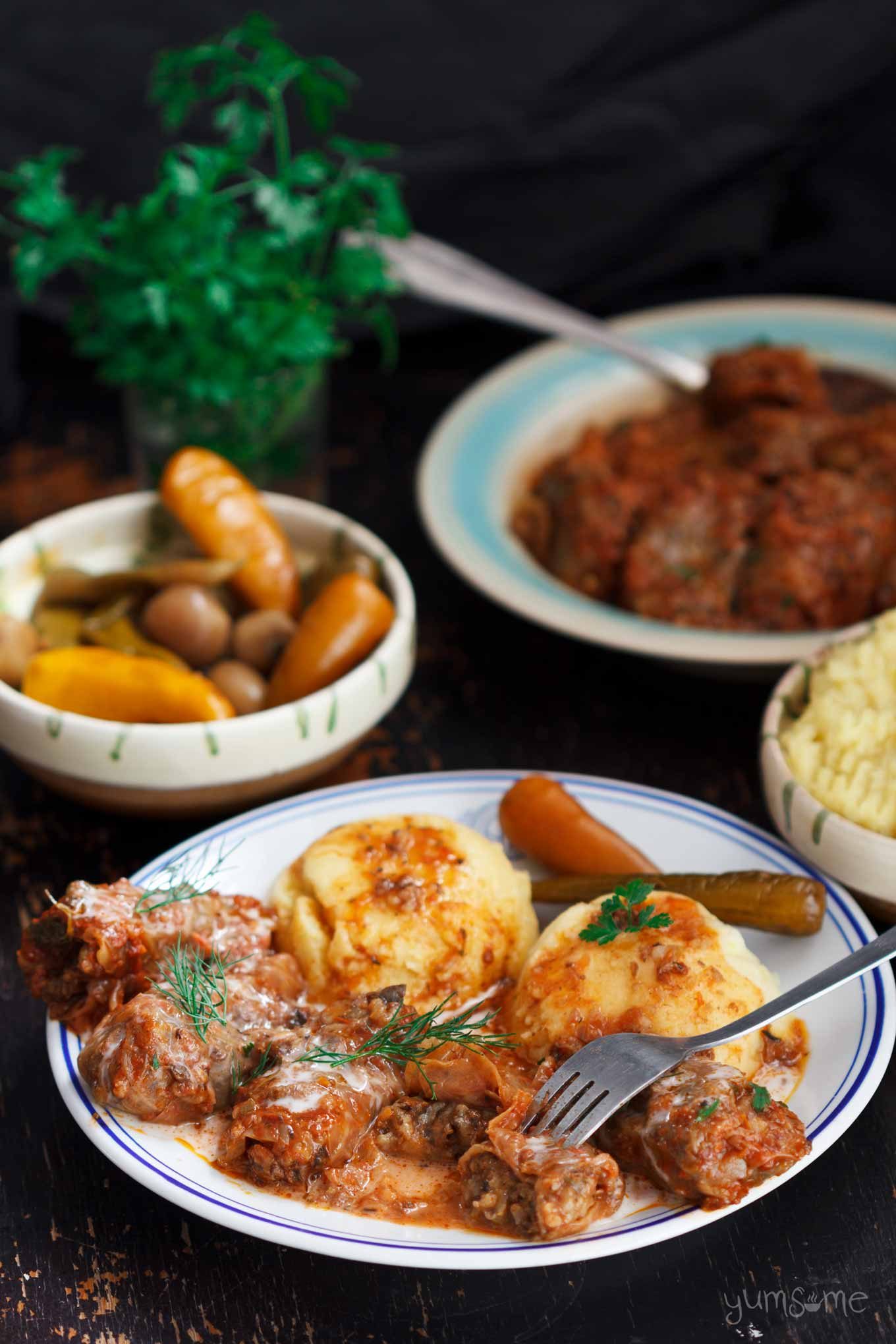 A black table laid with dishes of Romanian sarmale (cabbage rolls), mashed potatoes, and pickled chillies. In the background are more dishes of pickles and sarmale, plus fresh parsley.