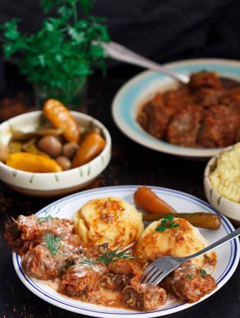 A black table laid with dishes of Romanian sarmale (cabbage rolls), mashed potatoes, and pickled chillies. In the background are more dishes of pickles and sarmale, plus fresh parsley.