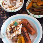 Closeup overhead shot of a blue and white dish of fasole with caramelised onions, sausages, and pickles. Dishes of fasole and pickles are in the background.