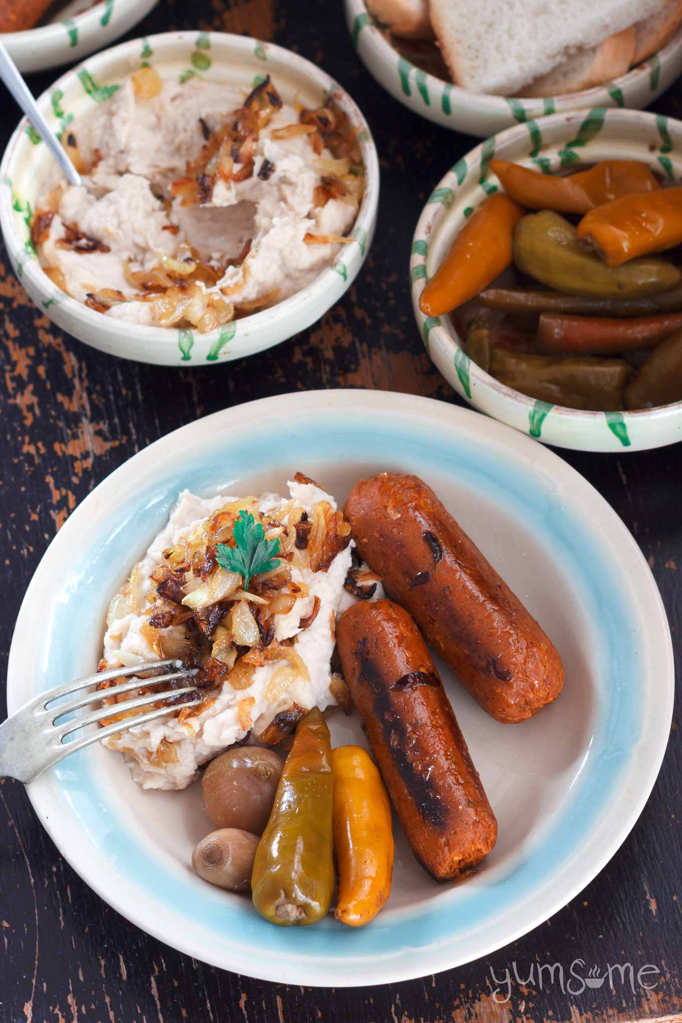 Closeup overhead shot of a blue and white dish of fasole with caramelised onions, sausages, and pickles. Dishes of fasole and pickles are in the background.