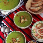 Overhead view of two bowls of piquant pea and leek soup on a red and white cloth, plus a toureen of soup and a basket of sliced bread.