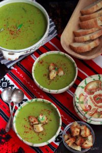 Overhead view of two bowls of piquant pea and leek soup on a red and white cloth, plus a toureen of soup and a basket of sliced bread.