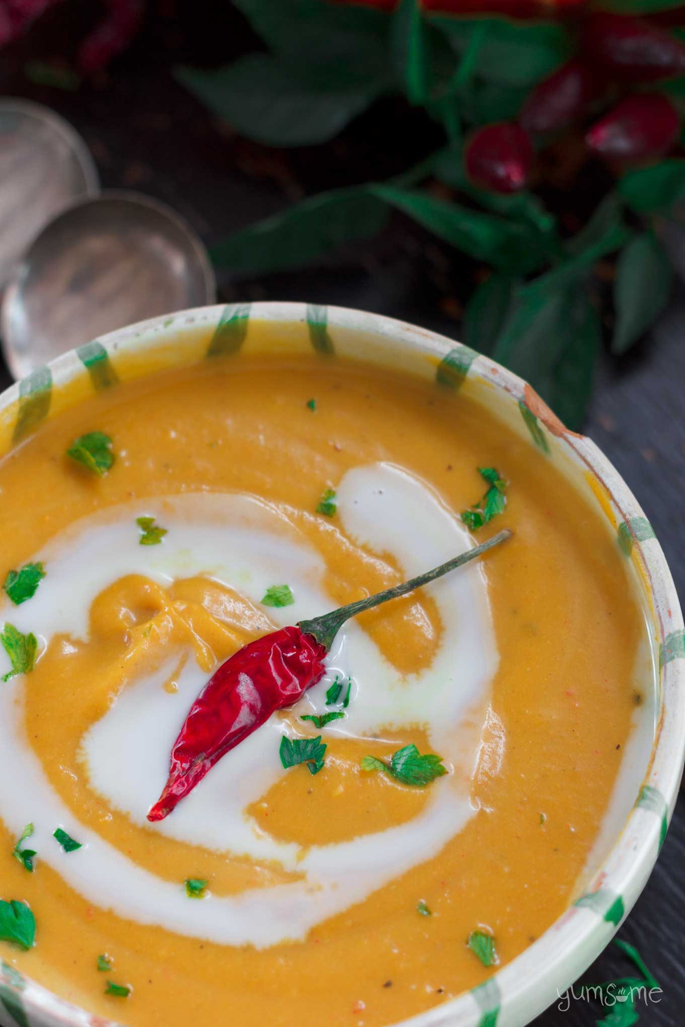 Overhead shot of a bowl of curried butternut squash soup with a swirl of coconut cream over the top. In the background are some green leaves and red chillies.