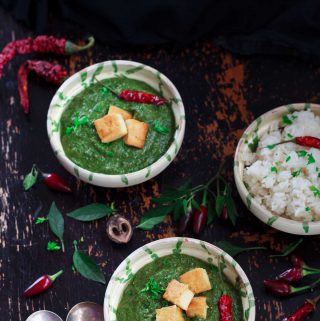 Overhead shot of two bowls of vegan palak paneer and some rice, surrounded by chillies on a dark background.