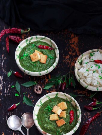 Overhead shot of two bowls of vegan palak paneer and some rice, surrounded by chillies on a dark background.