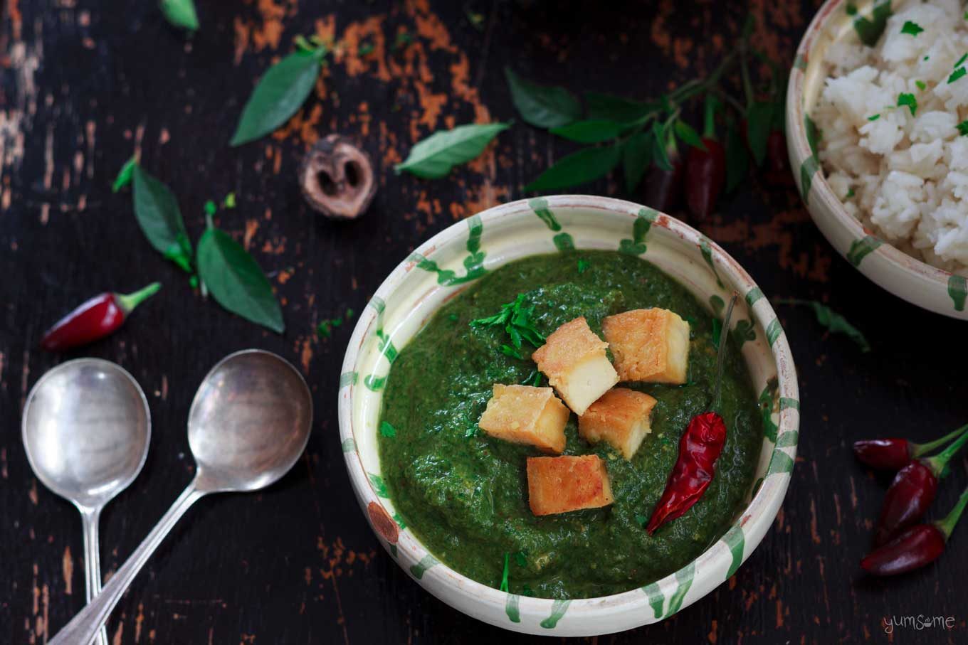 A bowl of vegan palak paneer and two silver spoons on a dark wood table.