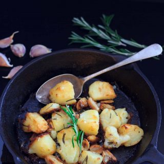 roast potatoes in a skillet with a serving spoon, some rosemary, and a few cloves of garlic | yumsome.com