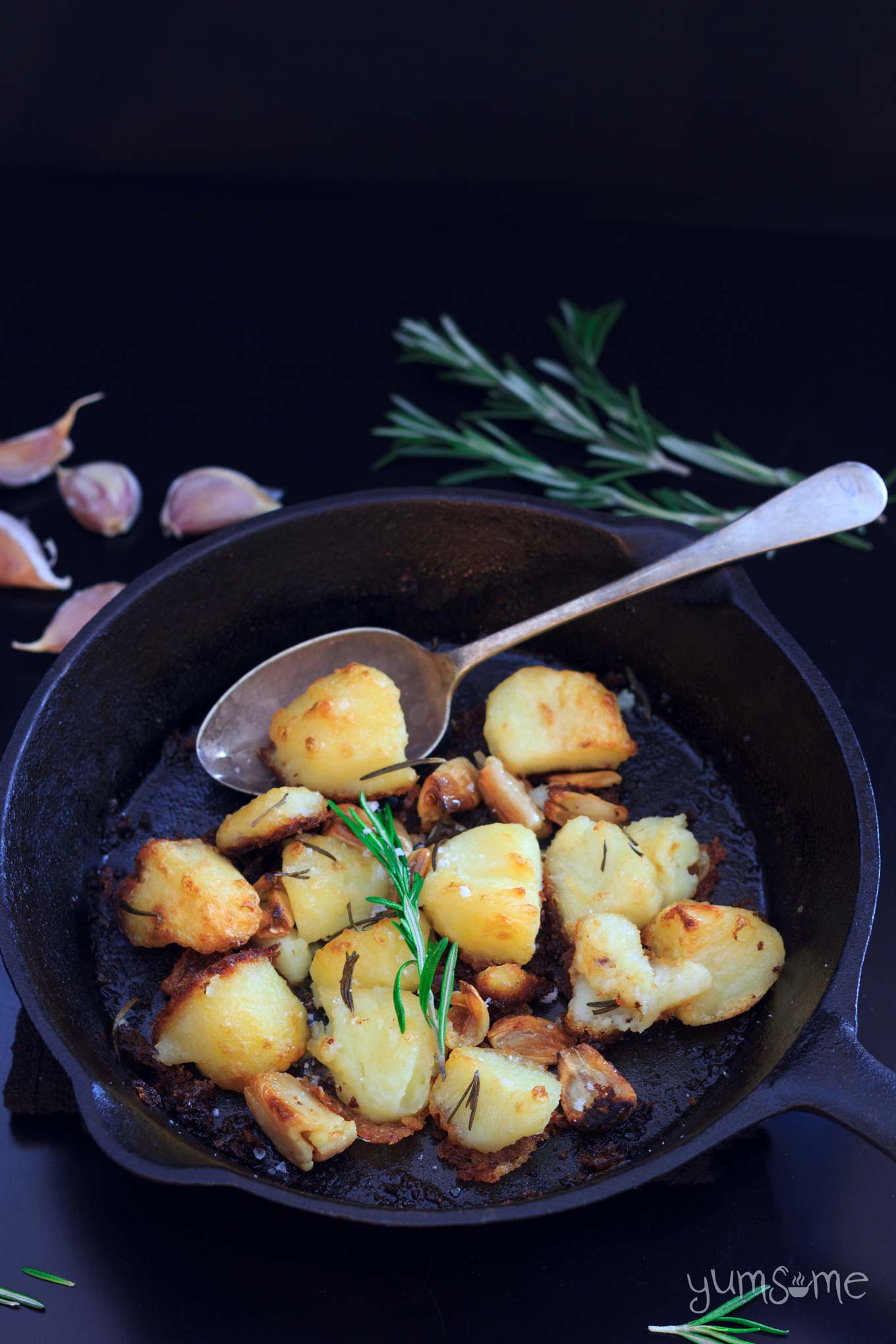 roast potatoes in a skillet with a serving spoon, some rosemary, and a few cloves of garlic | yumsome.com