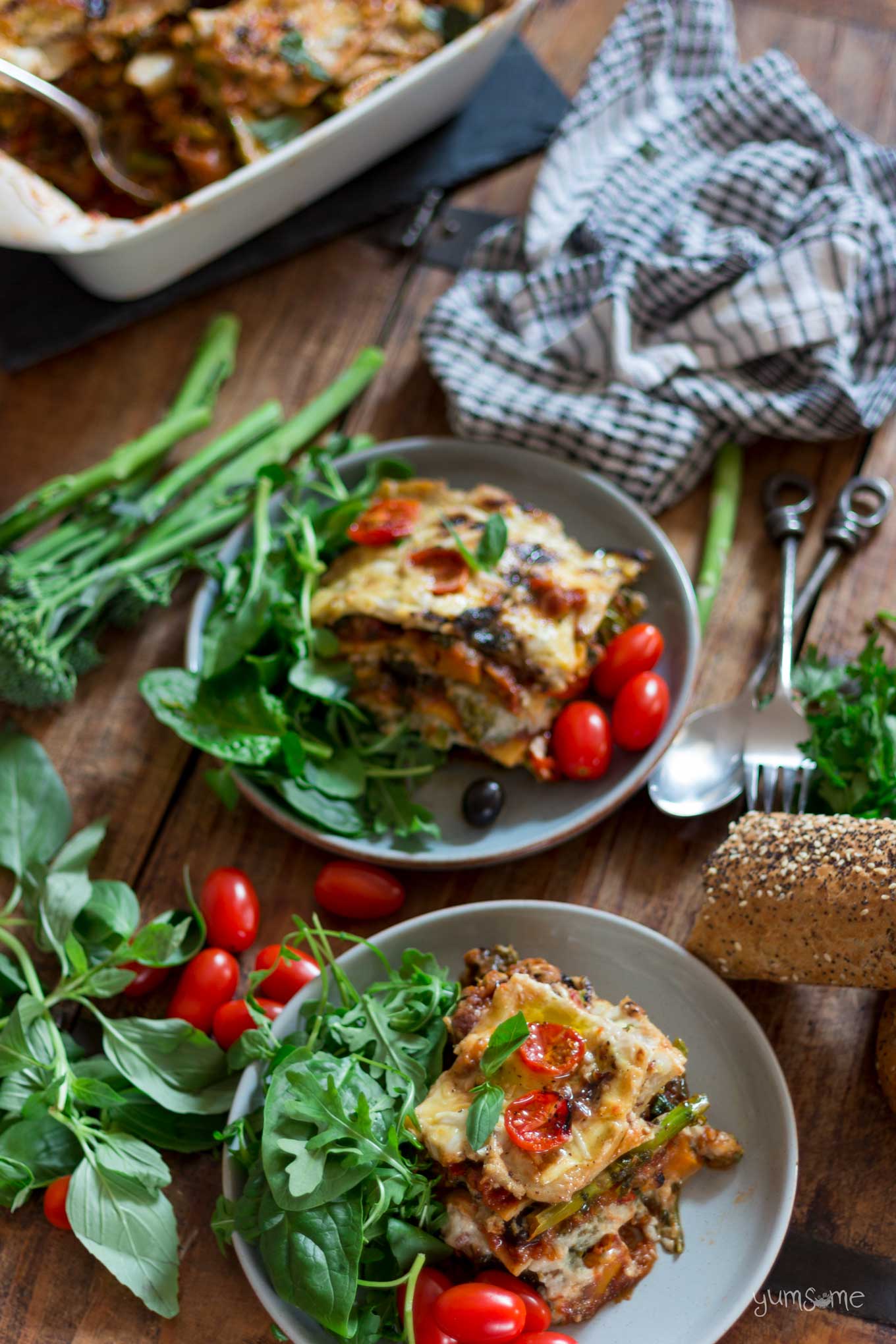 overhead shot of two plates of loaded vegan lasagne with green salad, baby plum tomatoes, tenderstem broccoli, basil, and some bread | yumsome.com