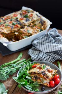 A large serving dish, plus a plate of loaded vegan lasagne, with green salad and tomatoes, plus a tea towel on a wooden table.