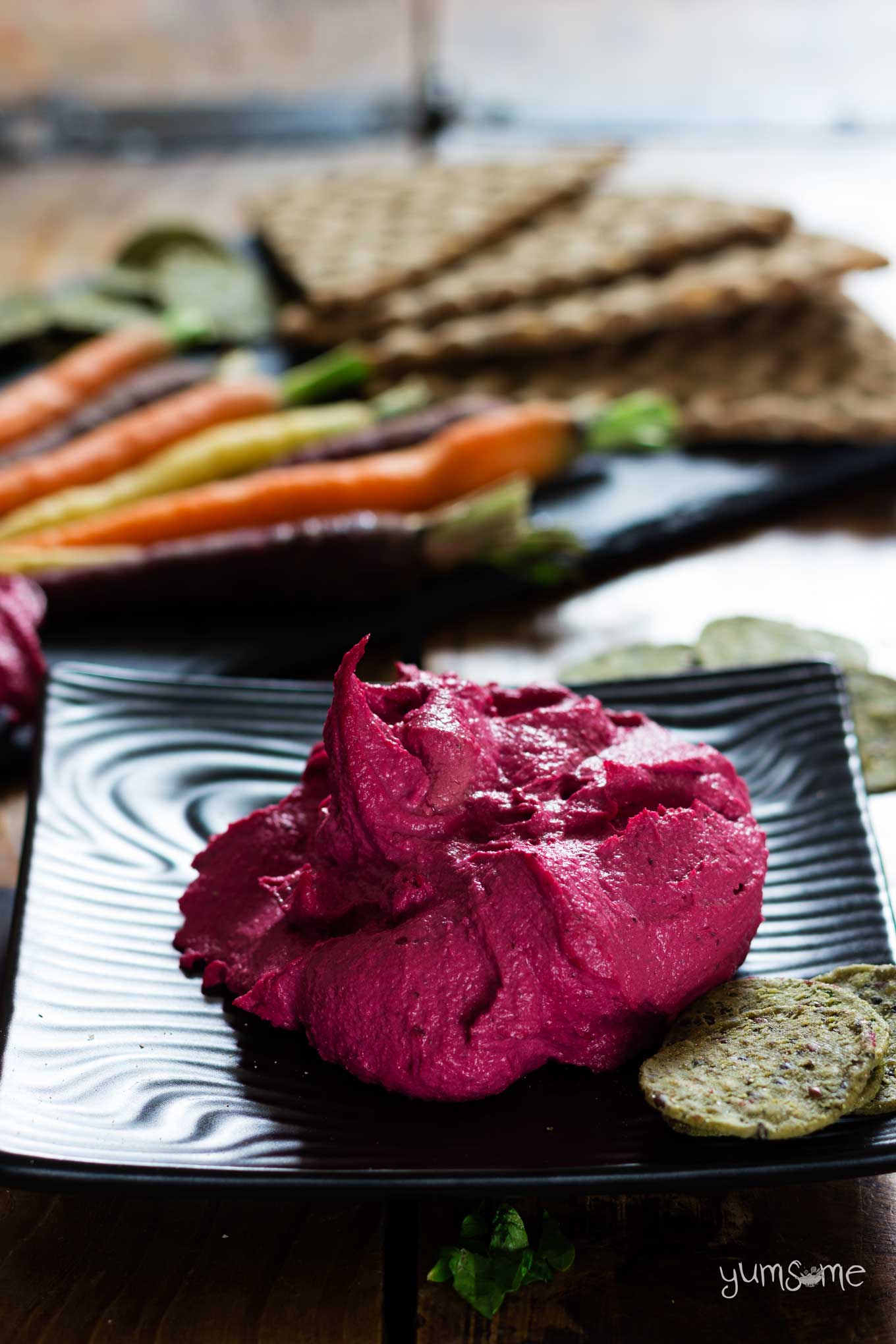 A pile of pink beetroot hummus on a black plate, with carrots in the background.