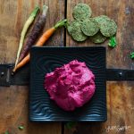 Green kale crackers, orange, purple, and yellow carrots, and a plate of pink beet hummus on a wooden table.