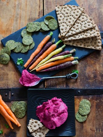 Bright pink beetroot hummus on a black plate, plus multicoloured carrots and crackers.