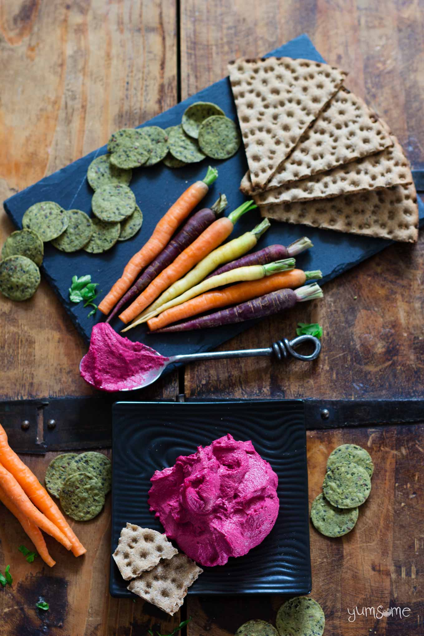 Bright pink beetroot hummus on a black plate, plus multicoloured carrots and crackers.