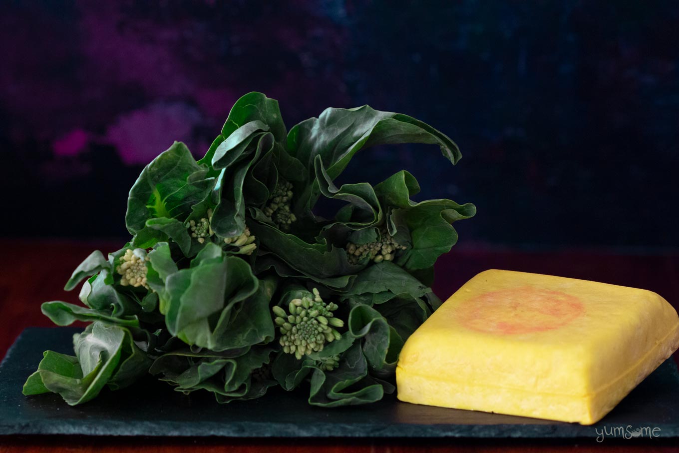 Chinese broccoli and a block of yellow tofu on a dark background.