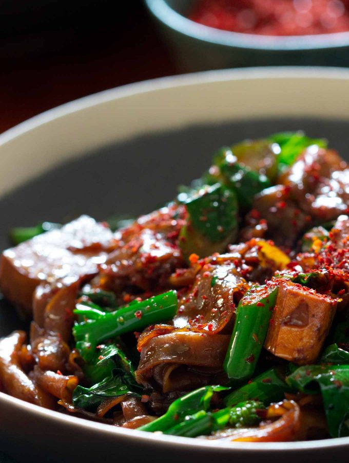 A dish of Chinese broccoli, fried rice noodles, and chunks of tofu, with a bowl of red chilli flakes in the background.