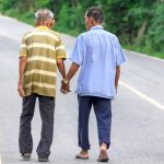 Two Thai men walking along a road, holding hands.