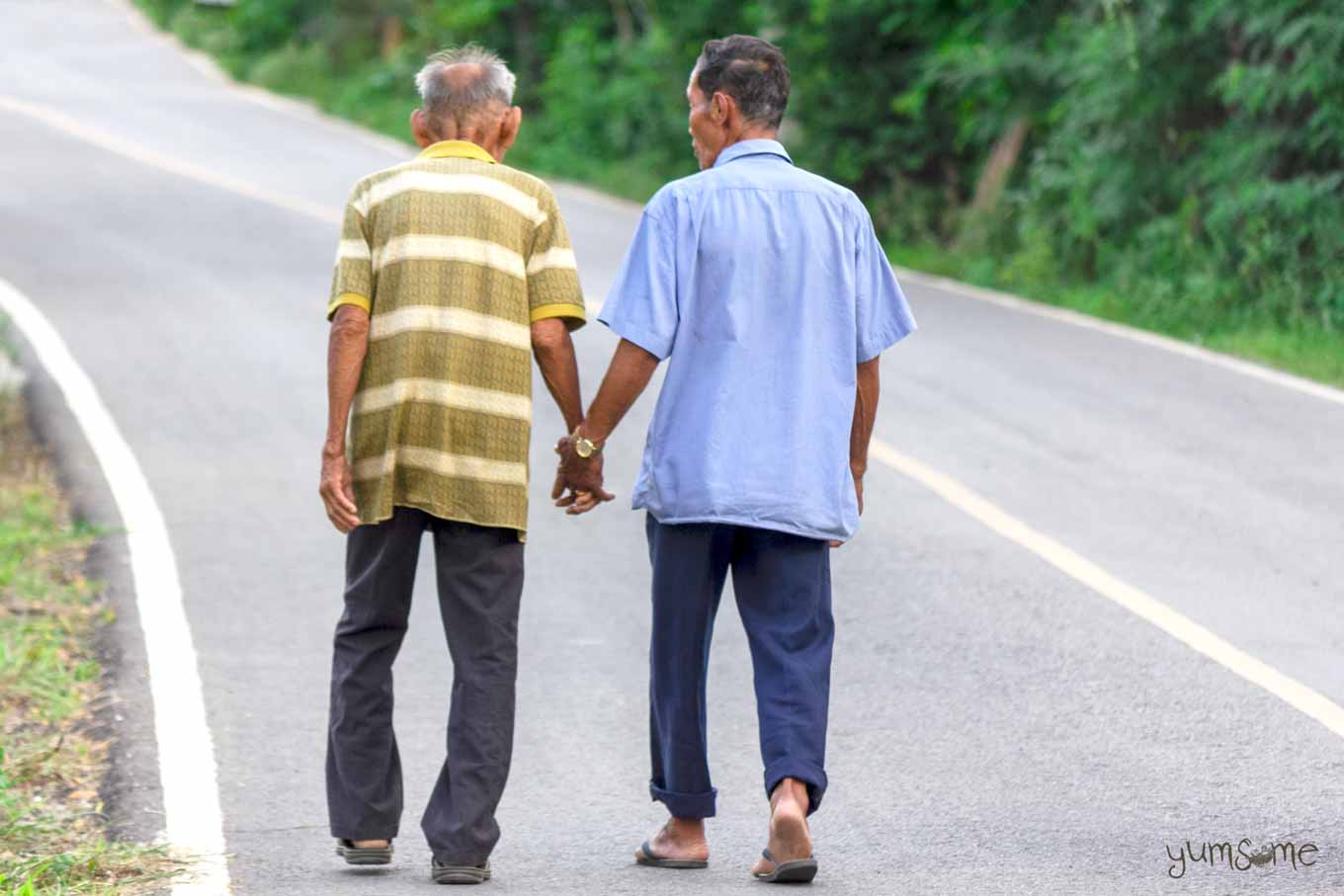 Two Thai men walking along a road, holding hands.