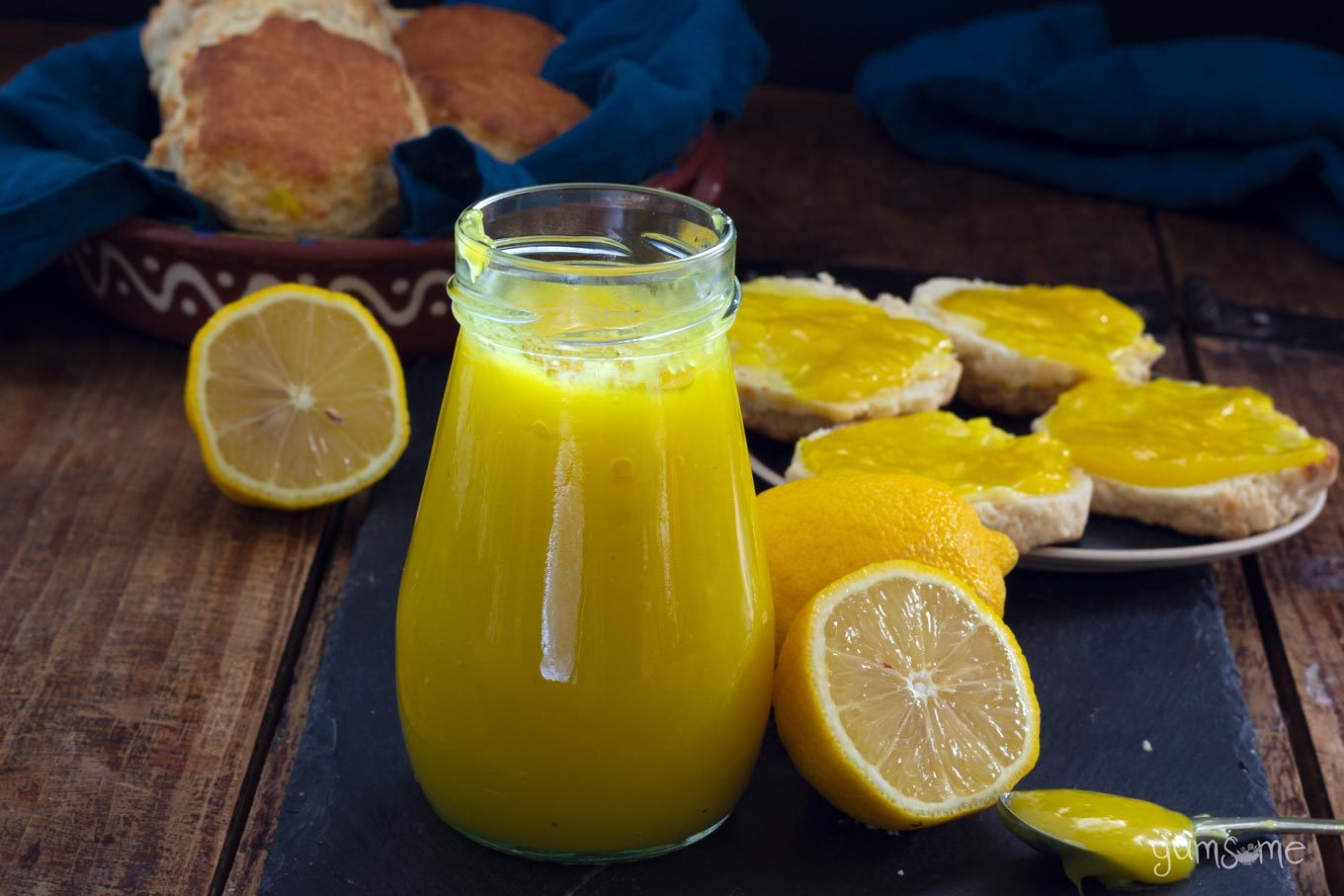 A jar of vegan lemon curd with some cut lemons, and scones spread with lemon curd in the background, all on a black slate on a wooden table.