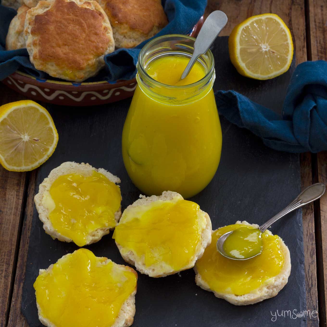 Some scones spread with lemon curd - one has a silver spoon resting on it. A jar of lemon curd, and more scones are in the background.