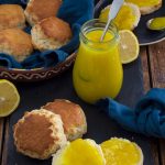 A bowl of whole scones, with some cut open and spread with lemon curd. A jar of lemon curd and some cut lemons are in the background.