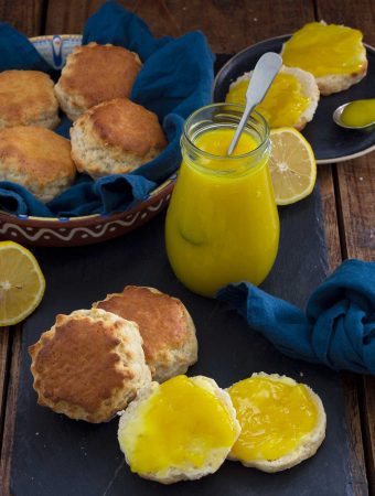 A bowl of whole scones, with some cut open and spread with lemon curd. A jar of lemon curd and some cut lemons are in the background.