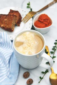 A white mug of vegan pumpkin spice latte against a white background, surrounded by spices, slices of cake, golden flatware, a pot of pumpkin puree, and a pale blue tea towel.