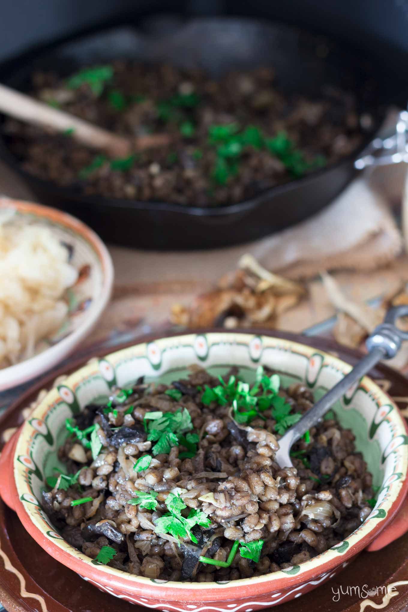 A fork sticks out of a dish of Czech mushroom and barley risotto. In the background is a black cast iron skillet containing more risotto, plus there's a plate of sauerkraut.