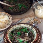 A wooden table with a dish of Czech mushroom and barley risotto, plus more risotto in a black skillet, along with a dish and a jar of sauerkraut.