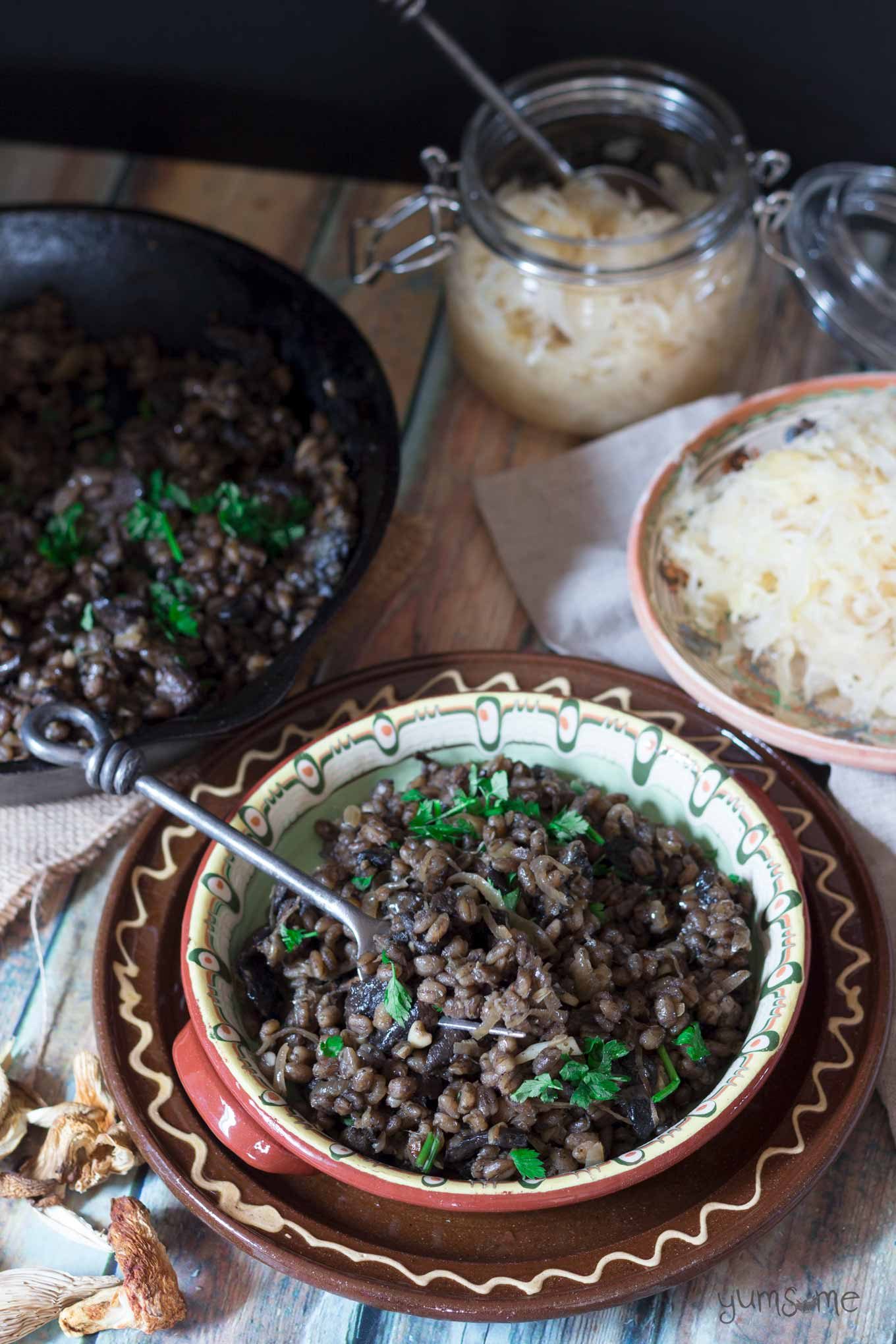 A rustic bowl of Czech mushroom barley risotto, with a cast iron skillet, and some sauerkraut on a plate in the background.