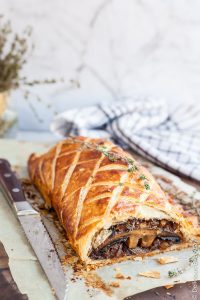 Vegan mushroom wellington on a wooden board, with a carving knife, and a pale blue tea towel.