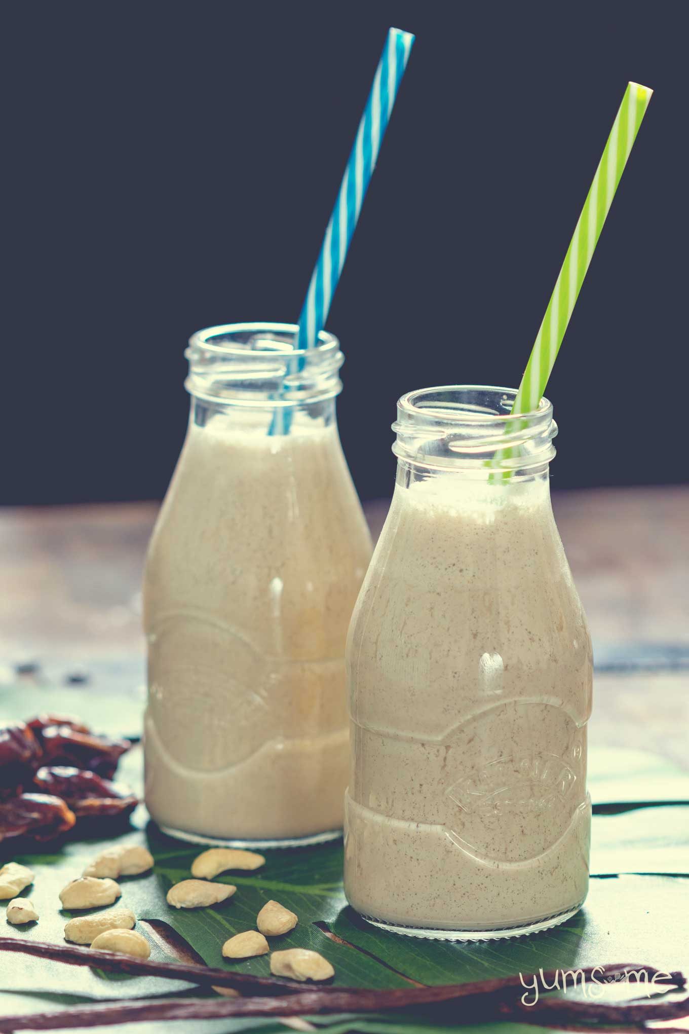 Two bottles of cashew milk with coloured straws, on a wooden table, surrounded by dates, cashews, and vanilla pods.