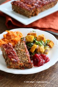 A white plate containing a slice of vegan lentil loaf, some roasted vegetables, and a blob of cranberry sauce. In the background is more lentil loaf on a white platter, and an orange napkin.